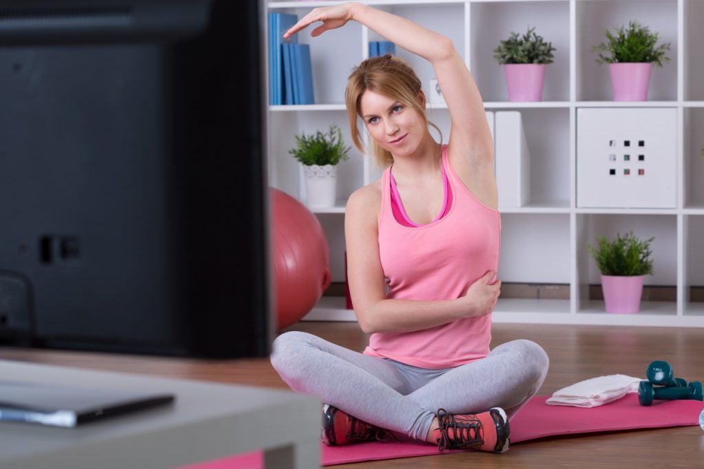 Mujer joven con ropa deportiva haciendo ejercicio frente a un televisor. Practicar deporte suave es beneficioso para aliviar el dolor de la fibromialgia reumática.
